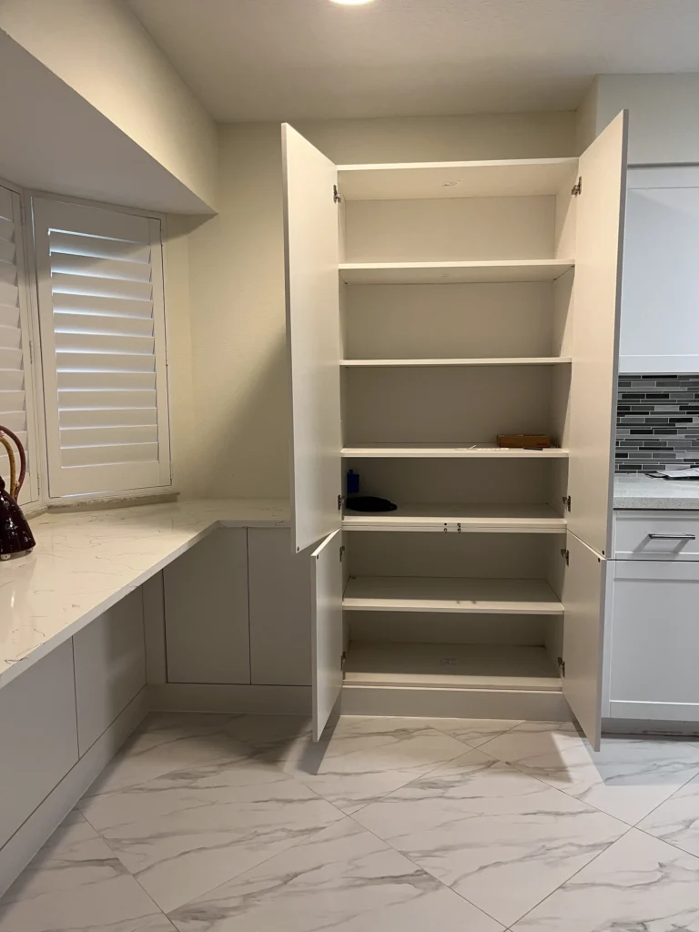 Tall pantry cabinet open to show adjustable shelves beside a quartz desk counter