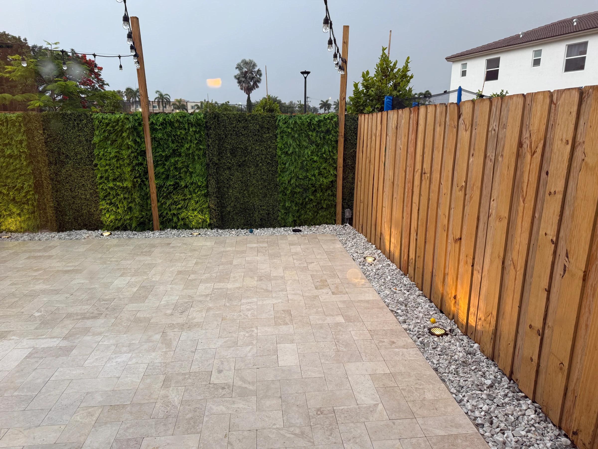New travertine-look paver patio with a gravel border, lit wood fence and a green hedge wall