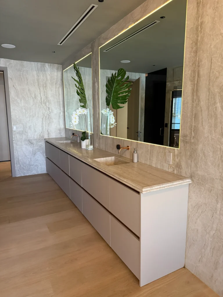 Long white flat-front vanity with a stone top running beneath two lit mirrors